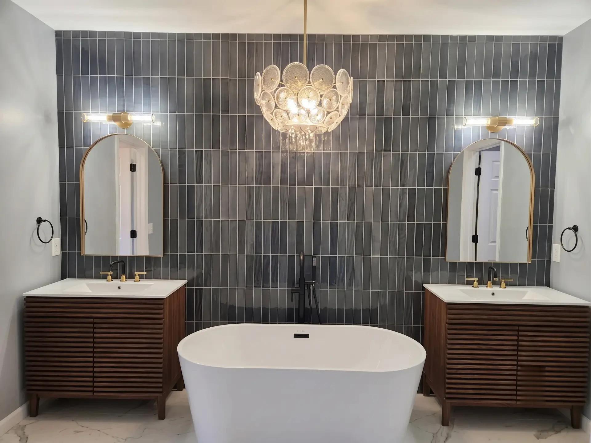 Symmetrical remodeled master bathroom with dark vertical tile, twin walnut vanities, arched mirrors and a freestanding soaking tub